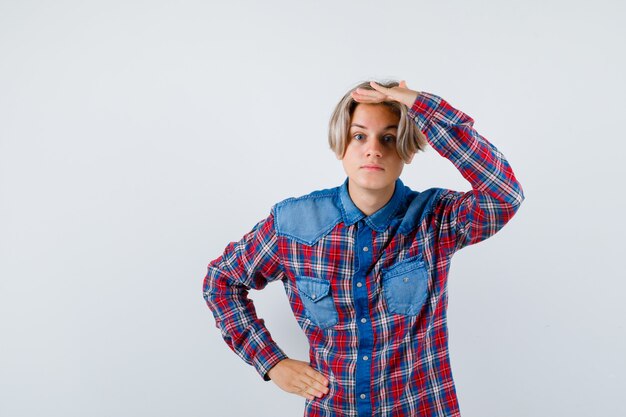 Foto portret van een jonge tienerjongen die de hand boven het hoofd houdt in een geruit overhemd en er nieuwsgierig uitziet aan de voorkant