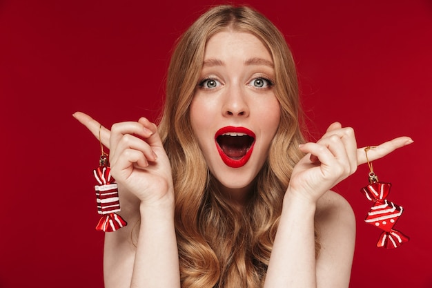 Portret van een jonge verraste blanke vrouw die zich voordeed op een rode muur met een felrode lippenstift die kerstsnoepspeelgoed vasthoudt.