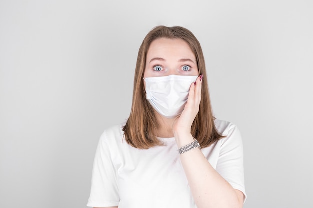 Portret van een jonge verraste vrouw in een medisch masker dat over grijze achtergrond wordt geïsoleerd