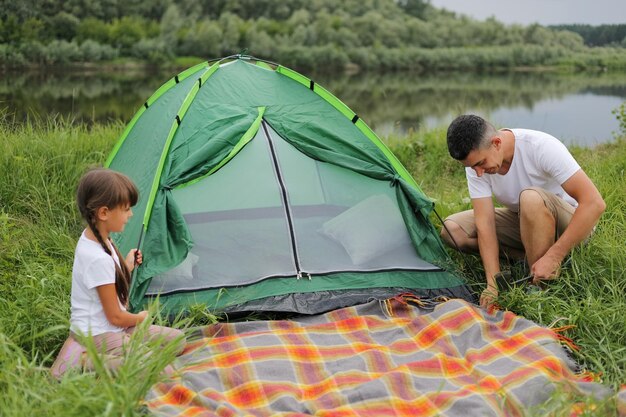 Portret van een jonge volwassen man met een wit t-shirt die een tent opzet in de natuur in de buurt van een meer- of rivierfamilie die samen op vakantie gaat picknicken in de open lucht en zich voorbereidt op een overnachting in de natuur