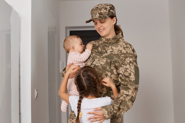 Portret van een jonge volwassen vrouwelijke soldaat die camouflage-uniform draagt, naar huis terugkeert nadat hij in het leger heeft gediend, een zelfverzekerde zorgzame moeder die haar kleine meisjes vasthoudt en knuffelt.