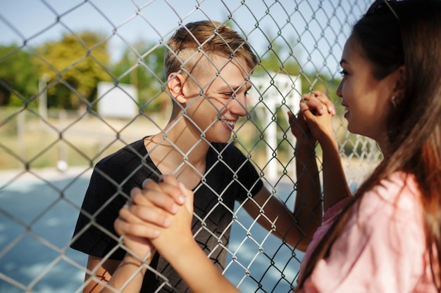 Portret van een jonge vrolijke jongen die op het basketbalveld staat terwijl hij vrolijk kijkt naar een aardig meisje met donker haar door een gaashek en haar hand vasthoudt