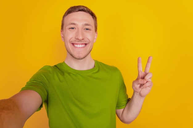 Portret van een jonge, vrolijke man die blij is met een positieve glimlach die een selfie neemt en een vredes cool v-teken toont, geïsoleerd over gele kleurachtergrond