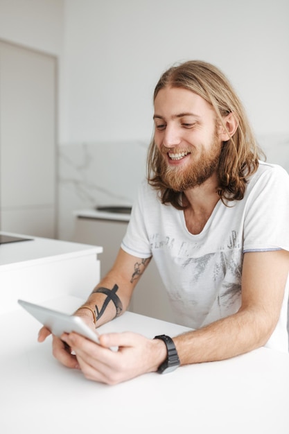 Portret van een jonge vrolijke man die thuis in de keuken zit en digitale tablet gebruikt