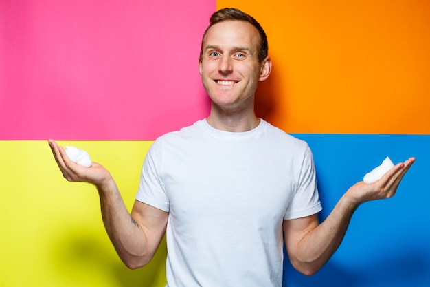 Portret van een jonge vrolijke man op een veelkleurige achtergrond, gekleed in een wit T-shirt, gegoten haarschuim in zijn handen, creëert een modieus kapsel