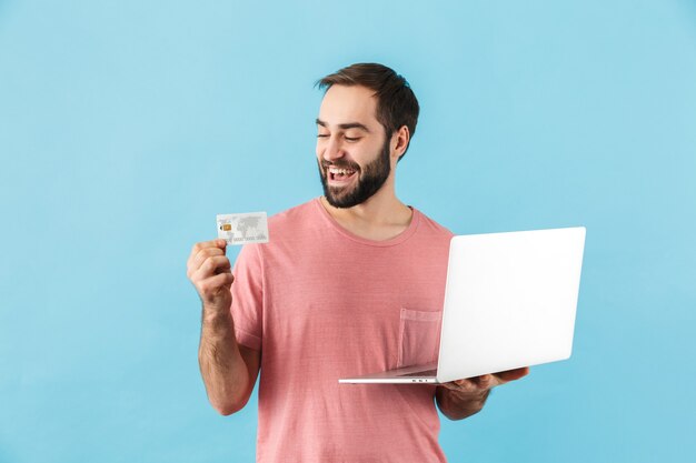 Portret van een jonge vrolijke opgewonden bebaarde man met een t-shirt dat geïsoleerd over een blauwe muur staat, een laptop vasthoudt en een plastic creditcard toont