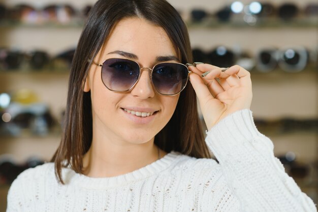 Portret van een jonge vrouw die, die zich in opslag winkelt en zonnebril dichtbij een spiegel probeert.