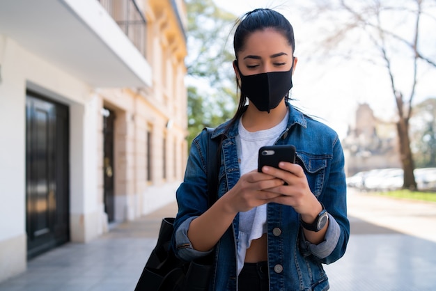 Portret van een jonge vrouw die een gezichtsmasker draagt en haar mobiele telefoon gebruikt tijdens het buiten lopen op straat. Nieuw normaal levensstijlconcept. Stedelijk concept.