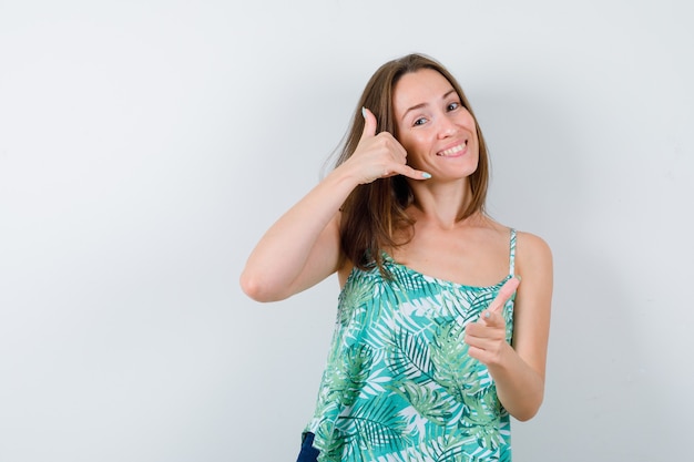 Portret van een jonge vrouw die een telefoongebaar toont, naar voren wijst en er vrolijk vooraanzicht uitziet