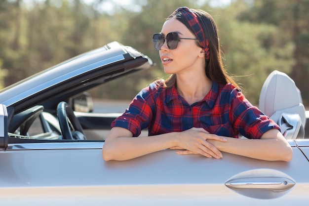 Portret van een jonge vrouw die een zonnebril draagt die in een cabrio zit en wegkijkt