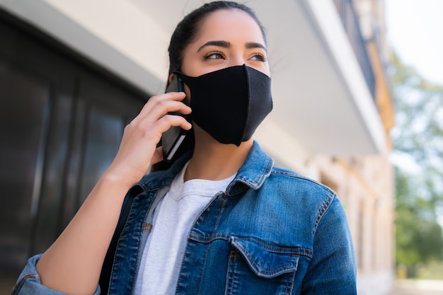 Portret van een jonge vrouw die gezichtsmasker draagt en aan de telefoon spreekt terwijl hij buiten op straat staat. Stedelijk concept