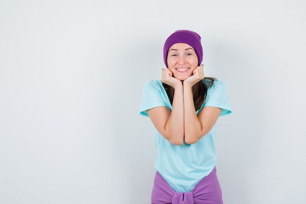 Portret van een jonge vrouw die haar gezicht op haar handen in t-shirt, beanie dempt en er vrolijk vooraanzicht uitziet