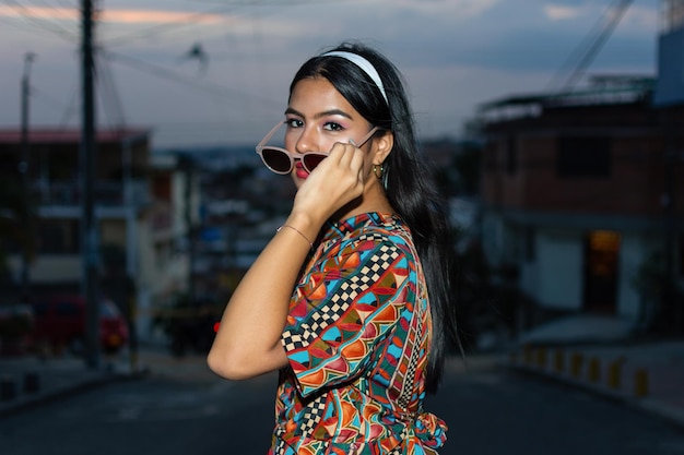Portret van een jonge vrouw die haar zonnebril vasthoudt met een kleurrijke jurk midden op straat en de stad op de achtergrond