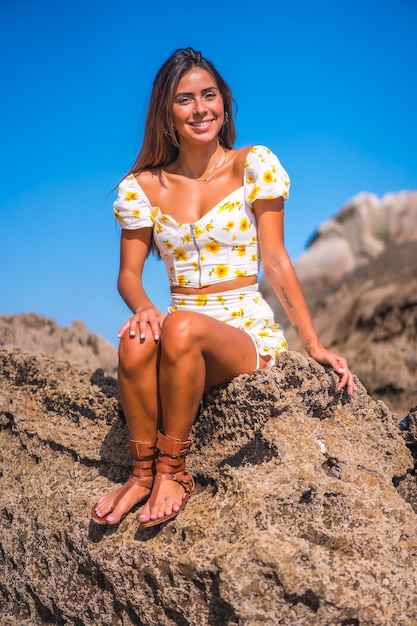 Portret van een jonge vrouw die lacht in een witte jurk op het strand, genietend van de zomer zittend op een paar rotsen