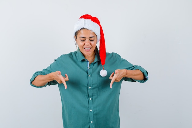 Portret van een jonge vrouw die naar beneden wijst in shirt, kerstmuts en er vrolijk vooraanzicht uitziet