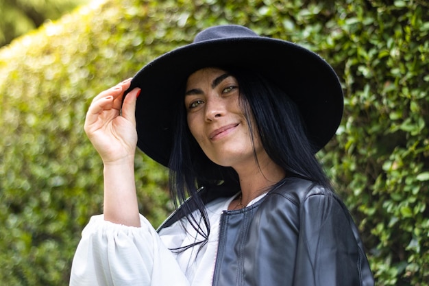 Portret van een jonge vrouw die naar de camera kijkt en haar hoed met groene bladeren op de achtergrond buiten vasthoudt