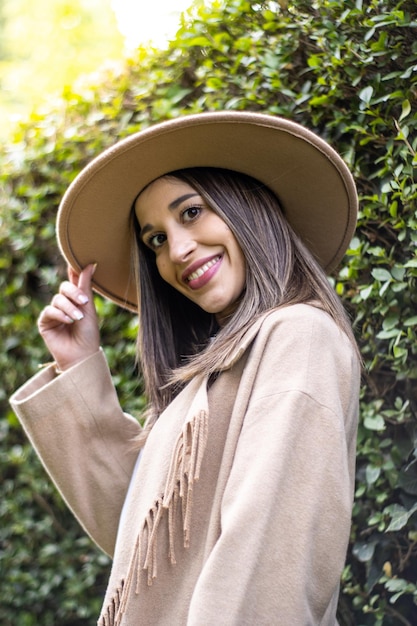 Portret van een jonge vrouw die naar de camera kijkt en haar hoed met groene bladeren op de achtergrond buiten vasthoudt