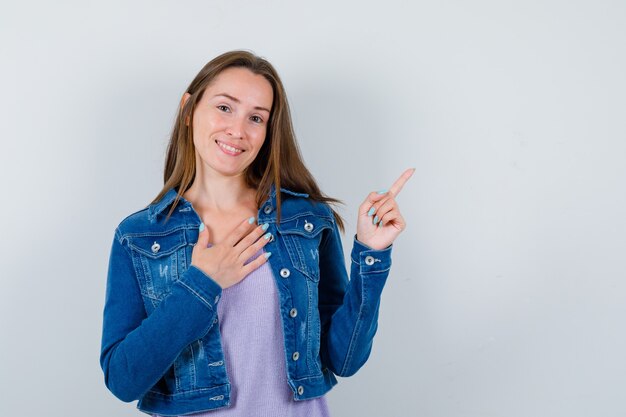 Portret van een jonge vrouw die naar de rechterbovenhoek wijst, met de hand op de borst in t-shirt, jas en dankbaar vooraanzicht