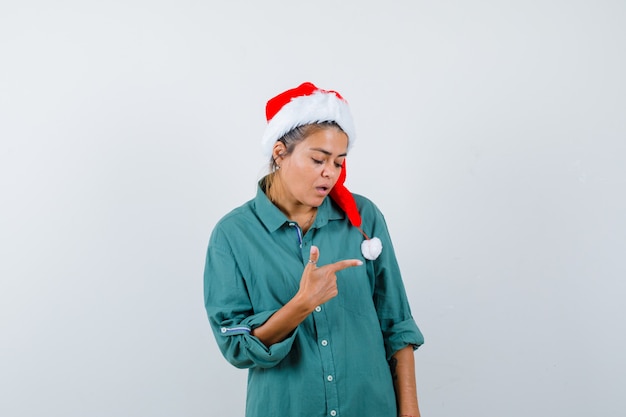 Portret van een jonge vrouw die naar de rechterkant wijst in shirt, kerstmuts en verbaasd vooraanzicht kijkt