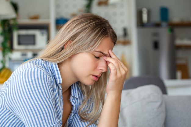 Portret van een jonge vrouw die thuis op de bank zit met hoofdpijn en pijn Mooie vrouw die lijdt aan chronische dagelijkse hoofdpijn Trieste vrouw die haar hoofd vasthoudt omdat sinuspijn