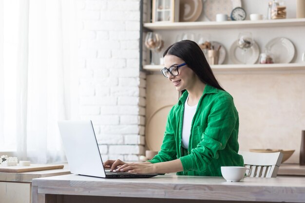 Portret van een jonge vrouw die thuis tijd doorbrengt in de keuken en een laptop gebruikt