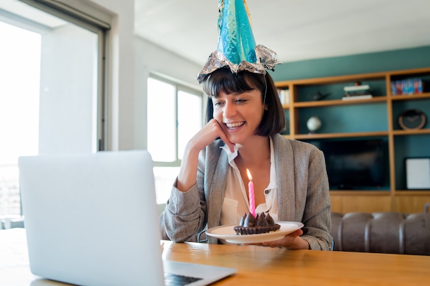 Portret van een jonge vrouw die verjaardag viert tijdens een videogesprek met laptop en cake van huis