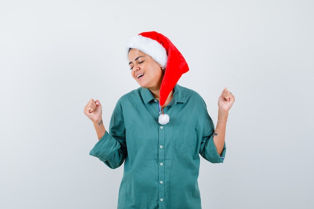 Portret van een jonge vrouw die winnaargebaar in overhemd, kerstmuts toont en opgetogen vooraanzicht kijkt