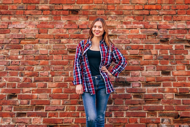 Portret van een jonge vrouw in een geruit overhemd en een spijkerbroek die tegen een rode bakstenen muur staat