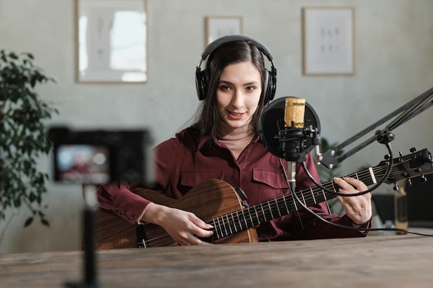 Portret van een jonge vrouw in een koptelefoon die gitaar speelt en een lied opneemt in de opnamestudio