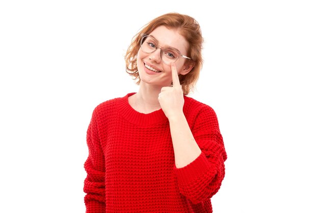 Portret van een jonge vrouw in een rode trui die met de vinger naar een bril op het gezicht wijst, geïsoleerd op een witte achtergrond van een studio.