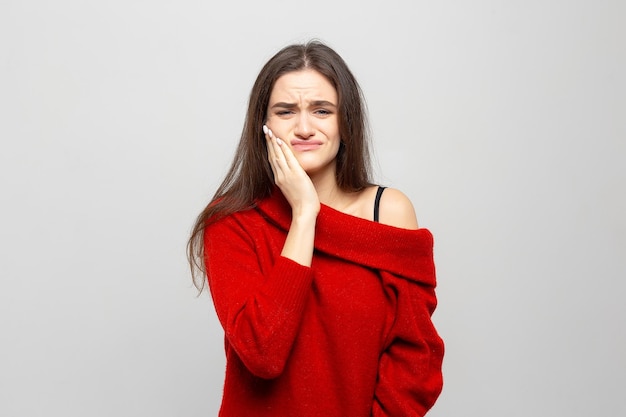 Portret van een jonge vrouw in een rode trui met ernstige kiespijn houdt haar hand op haar wang op een grijswitte achtergrond