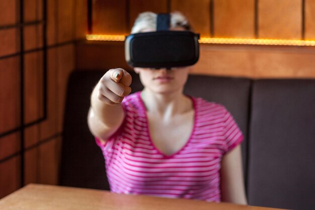 Portret van een jonge vrouw in een roze t-shirt die in een café zit, een virtual reality-headset draagt en video kijkt, een spel speelt op de simulator en met de vinger naar de camera wijst. Binnen, levensstijl