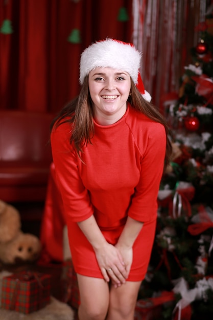 Foto portret van een jonge vrouw in helper kerstmuts en rode jurk ter voorbereiding op kerstmis thuis. kerstmis, kerstmis, winter, geluksconcept - glimlachende vrouw op de boommuur van de kerstmisglans.