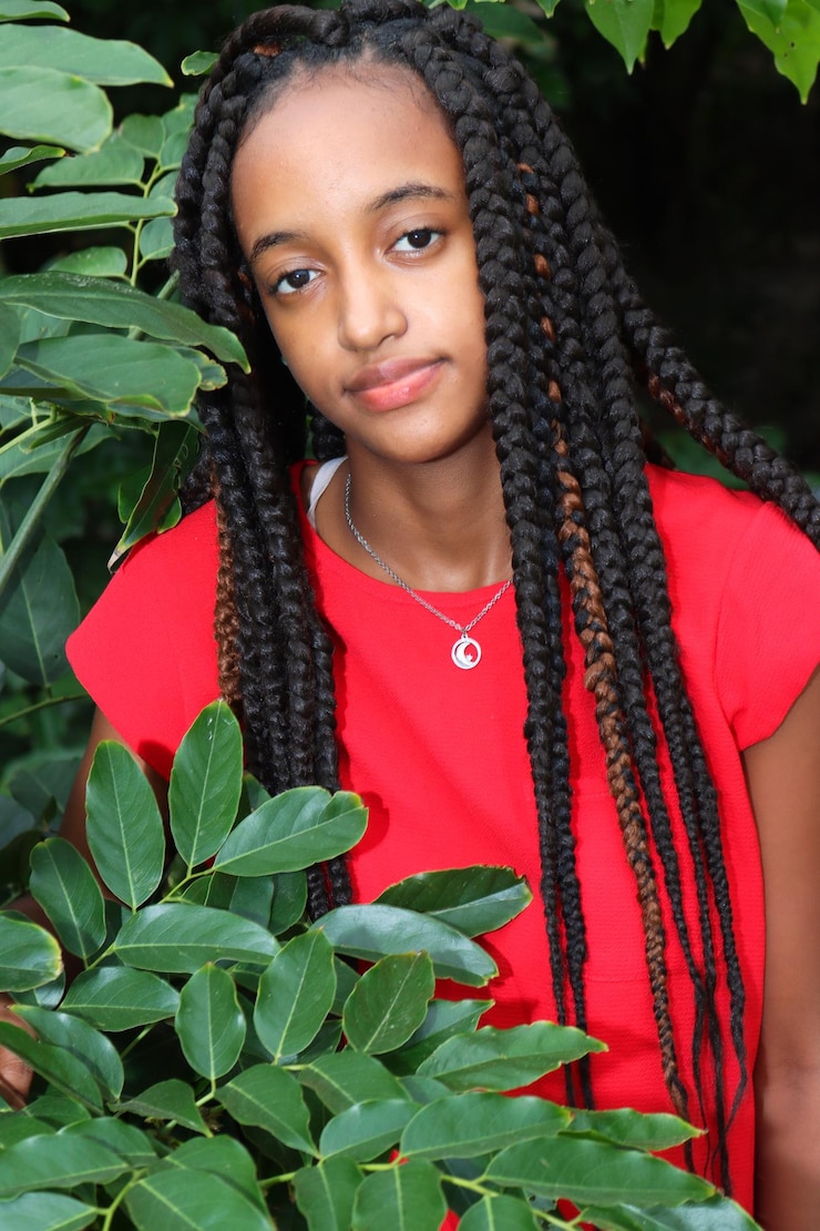 Foto portret van een jonge vrouw in het rood met lange vlechten die tegen planten staat