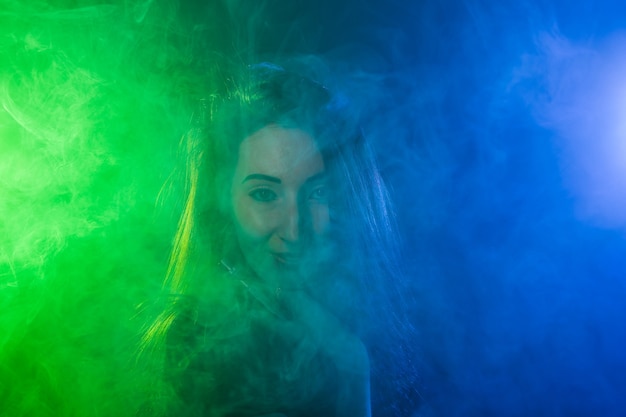 Portret van een jonge vrouw in neon blauwe en groene rook met vape of e-sigaretten.