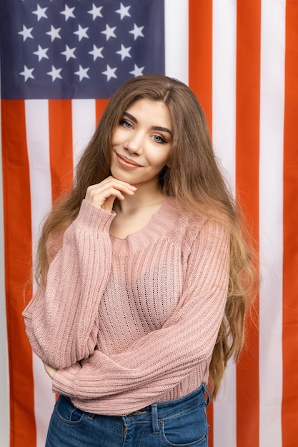 Foto portret van een jonge vrouw in spijkerbroek en een trui die tegen de achtergrond van de amerikaanse vlag in de camera kijkt het concept liefde voor het moederland