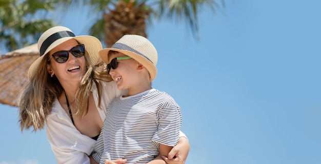 Portret van een jonge vrouw in strandkleding die haar zoon omhelst Moeder en kind genieten van zomervakantie in tropisch land met palmbomen op de achtergrond kopie ruimte