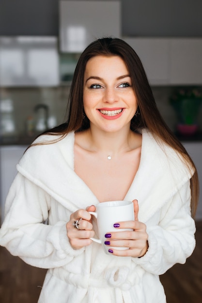 Portret van een jonge vrouw in wit gewaad na bad houdt een kopje koffie of thee tegen keuken achtergrond. Close-up gezicht brede glimlach.