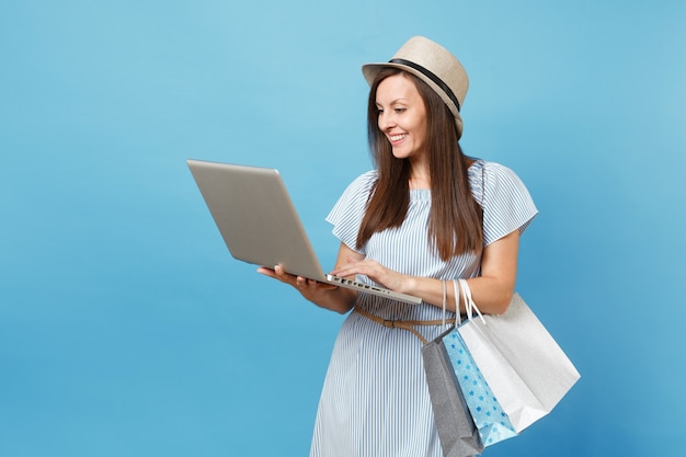 Portret van een jonge vrouw in zomerjurk, strohoed met pakketten met aankopen na online winkelen, met behulp van laptop pc-computer geïsoleerd op blauwe pastelachtergrond. ruimte voor advertentie kopiëren