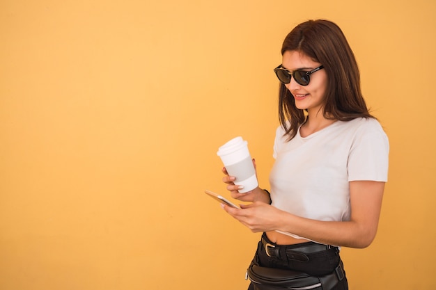 Portret van een jonge vrouw met behulp van haar mobiele telefoon terwijl ze een kopje koffie buiten op straat houdt