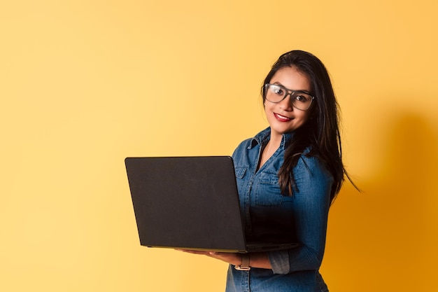 Portret van een jonge vrouw met een bril die een laptop vasthoudt en glimlachend naar de camera kijkt op een gele achtergrond