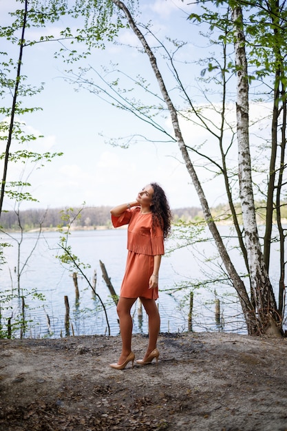 Portret van een jonge vrouw met een glimlach op haar gezicht, gelukkige emoties. Een meisje met lang golvend krullend haar in een jurk in de zomer op de natuur, in een bos aan een meer, stond in de buurt van een weg en struiken