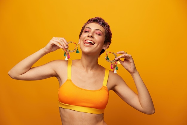 Portret van een jonge vrouw met een kort kapsel en gekleurd haar glimlachend en haar tong tonend naar de camera op een oranje achtergrond met oorbellenaccessoires in de studio