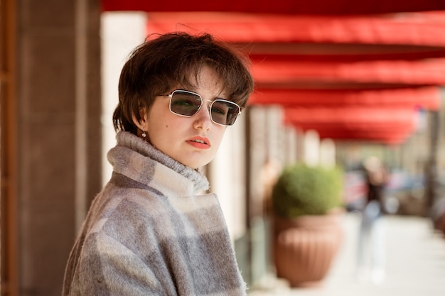 Portret van een jonge vrouw met een kort kapsel in een grijze jas en zonnebril op een straat in de stad op een zonnige lentedag