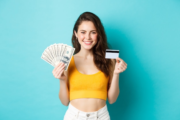 Portret van een jonge vrouw met een lachend gezicht, met plastic creditcard en geld, staande tegen een blauwe achtergrond.