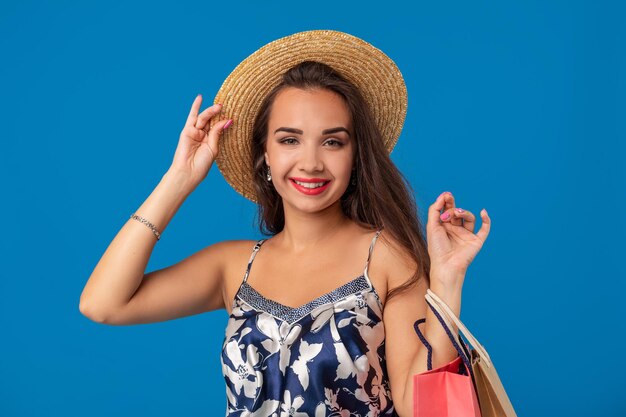 Portret van een jonge vrouw met een zomerhoed die boodschappentassen vasthoudt en naar de camera kijkt die over een blauwe achtergrond wordt geïsoleerd