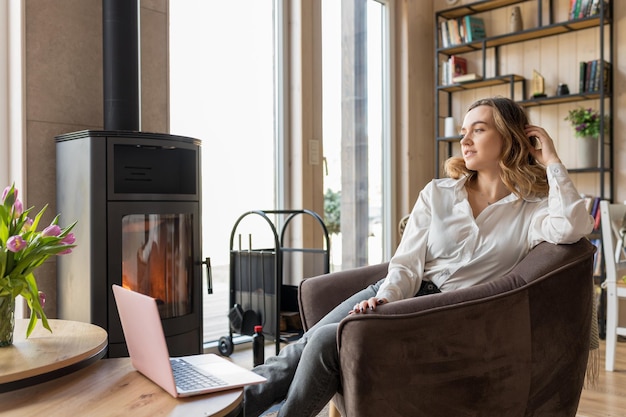 Portret van een jonge vrouw met laptop zittend op een zachte fauteuil in casual kleding in de buurt van raam en firep