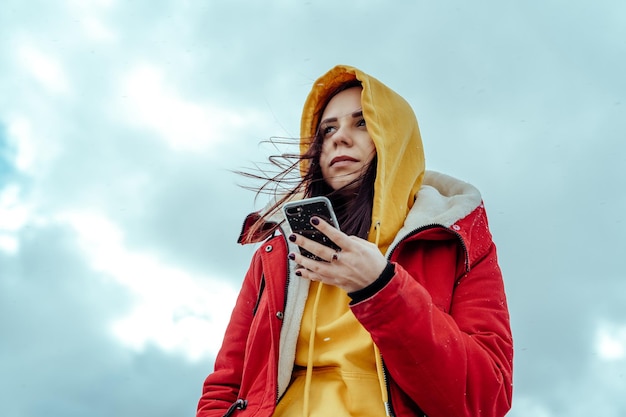 Portret van een jonge vrouw met mobiele telefoon op de achtergrond van de hemel Brunette in gele hoodie en rode jas die smartphone doorbladert bij bewolkt weer