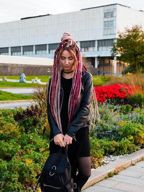 Portret van een jonge vrouw met roze dreadlocks in zwarte kleding met een rugzak die in het park poseert