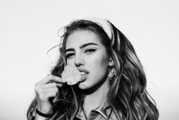 Portret van een jonge vrouw, mooie lippen, lichte make-up vasthouden, gebakken aardappel, friet, chips eten en poseren op blauwe achtergrond. Ongezond eten. Mooie vrouw die chips eet, sluit omhoog.